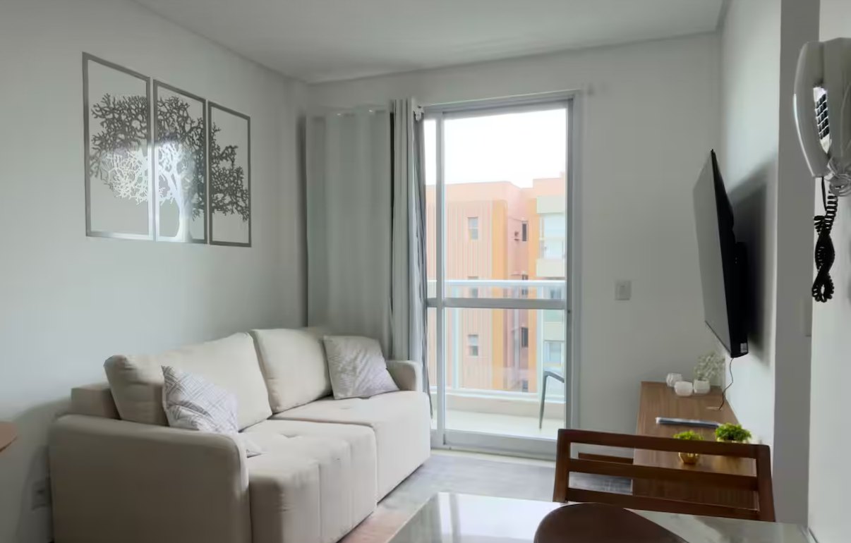 Sala clara com sofá bege, quadro de árvore na parede e porta de vidro para a varanda