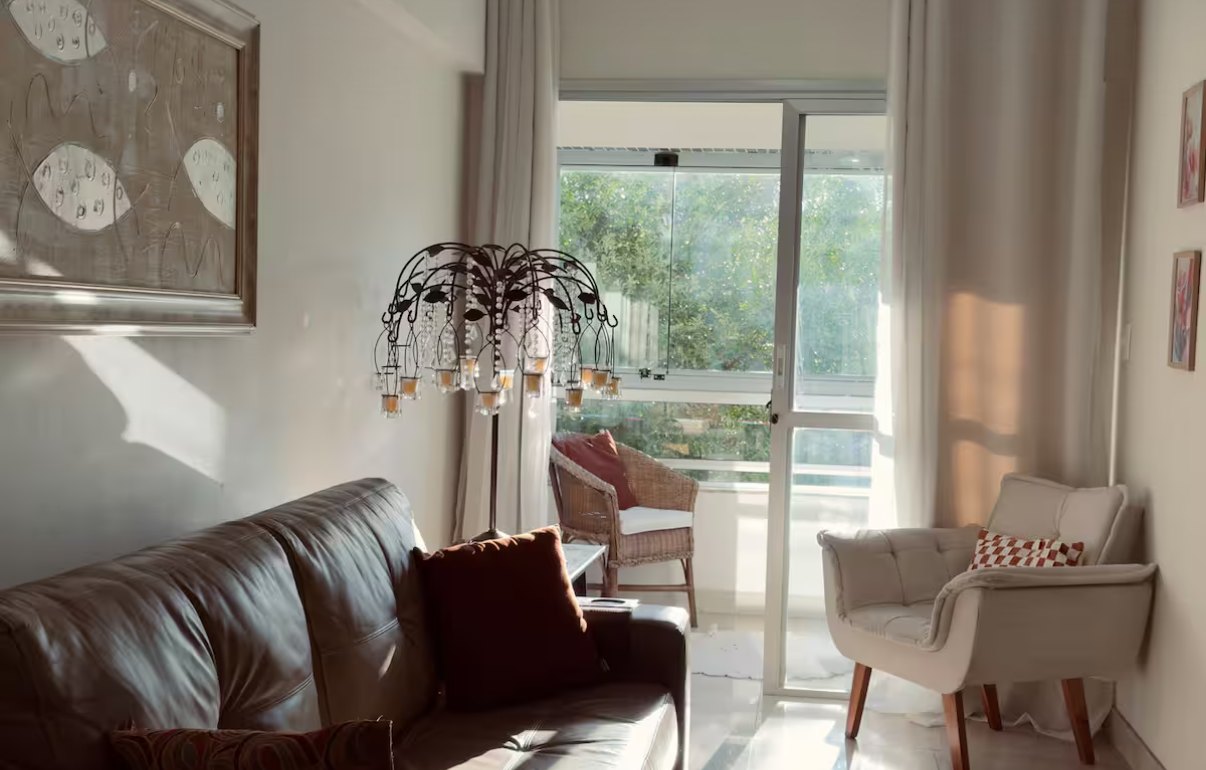 Sala aconchegante com sofá de couro, poltrona clara e varanda com cadeira de vime ao fundo