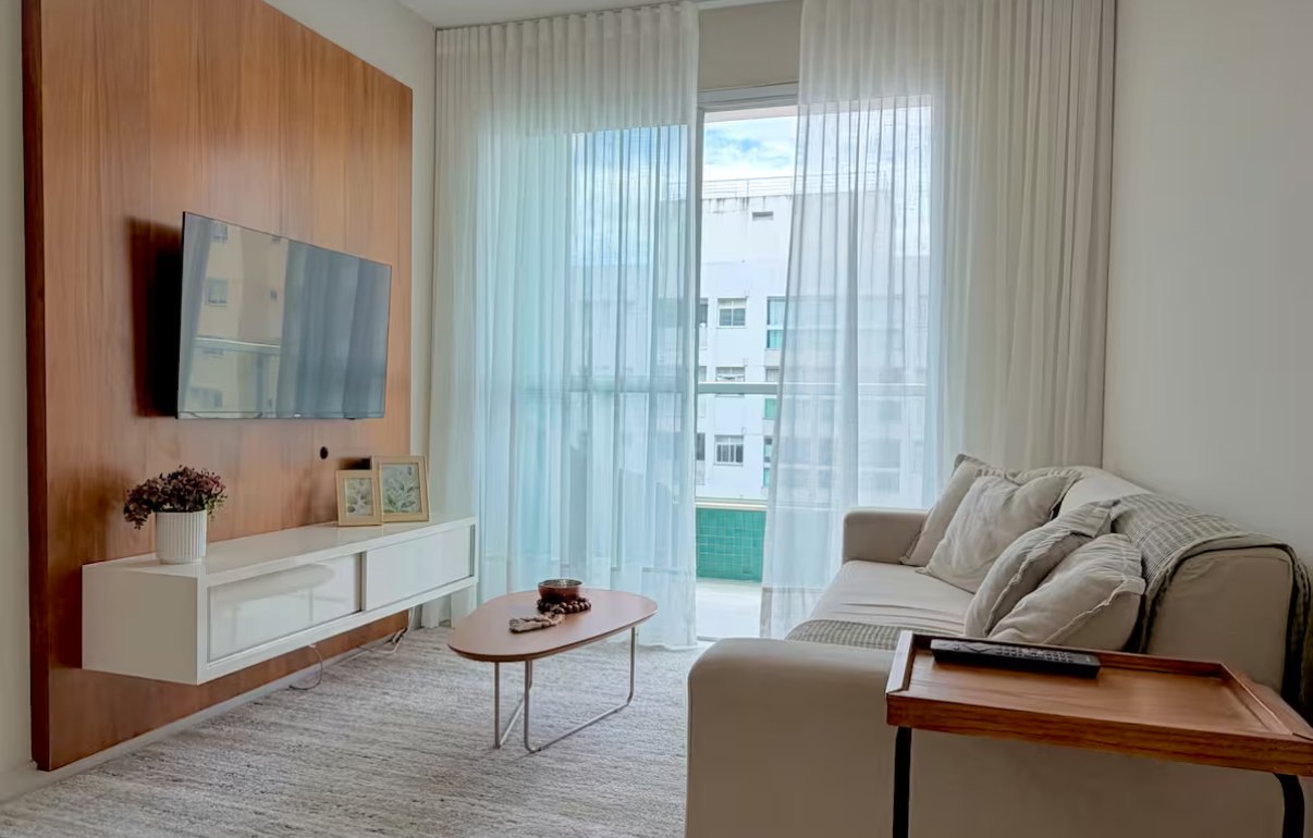 Sala com painel de madeira, televisão, sofá claro e cortinas leves na porta da varanda