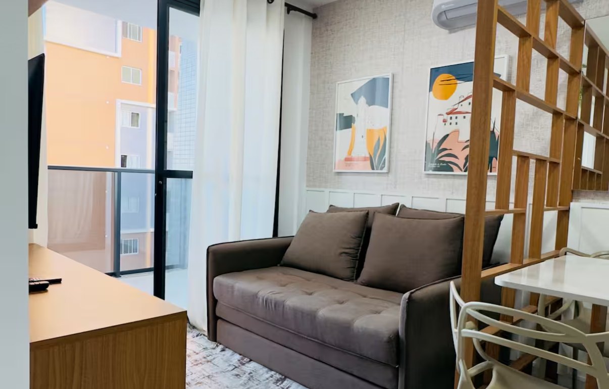 Sala com sofá marrom, quadros coloridos, divisória de madeira e porta de vidro para a varanda