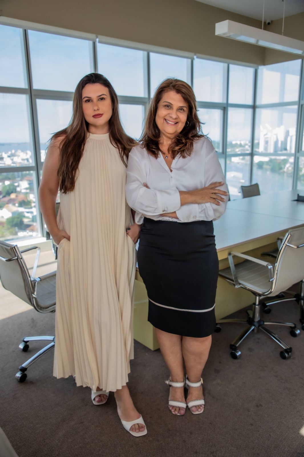Camila e Rita em pé em uma sala de reuniões com janelas amplas e vista para a cidade