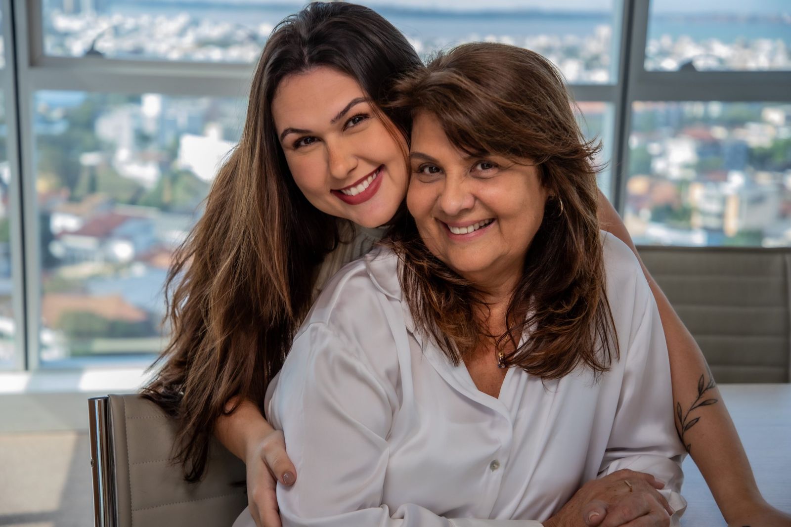 Camila e Rita, filha e mãe, abraçadas e sorrindo, com a vista da cidade de Vitória ao fundo