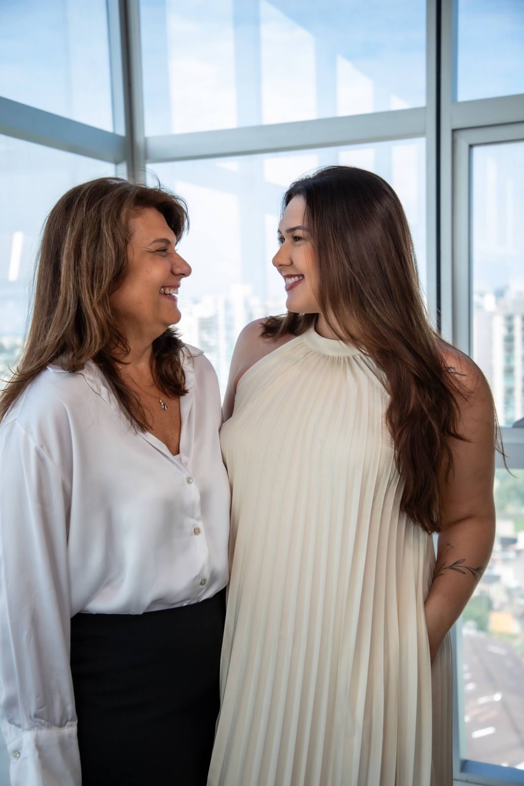 Rita e Camila sorrindo uma para a outra diante de uma janela ampla com vista da cidade